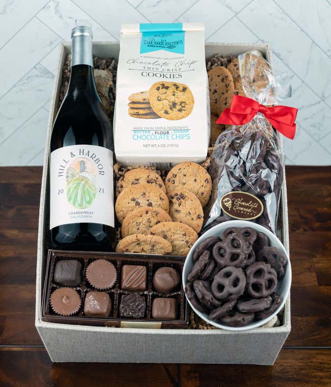a bottle of white wine, two glasses of the wine, a bag of milk chocolate pretzel balls, a box of chocolate chip cookies, a box of assorted chocolates, and a cutting board all alid out on a white wooden table