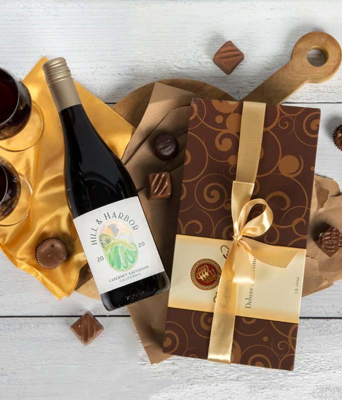 a bottle of red wine and a box of assorted chocolates laid out on a table with glasses of wine, cloth napkins, loose chocolates, and a cutting board