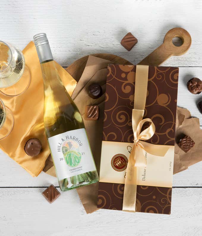 a bottle of white wine and a box of assorted chocolates laid out on a table with glasses of wine, cloth napkins, loose chocolates, and a cutting board
