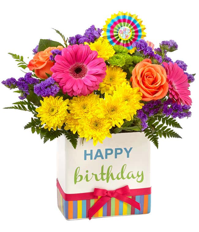Orange roses, pink gerbera daisies, yellow mums, purple filler, and fresh floral greens with a birthday pick, in a cube vase that says happy birthday