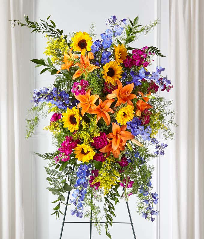 Standing spray or orange lilies, sunflowers, blue delphinium, fuchsia stock, and fresh floral greens 