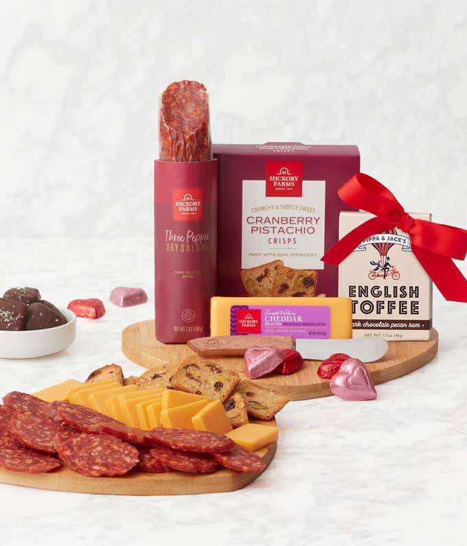 A serving board with a stick of salami, block of cheddar, boxes of cranberry pistachio crisps and english toffee, cheese spreader, and foil-wrapped chocolate hearts. 