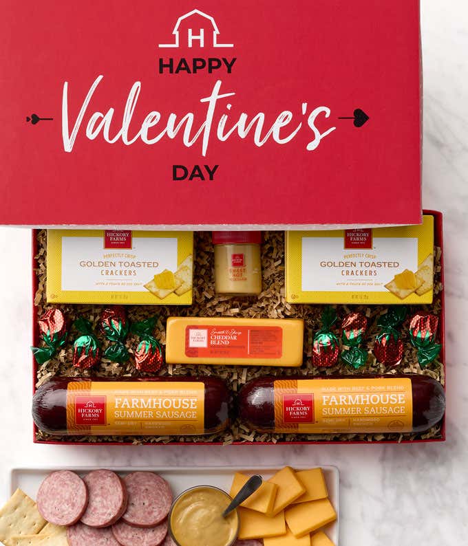 A "Happy Valentine's Day Gift Box with two boxes of golden toasted crackers, a jar of sweet hot mustard, one block of cheddar cheese, two  summer sausages, and strawberry bonbons laid out inside the gift box 