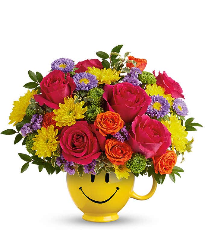 Hot Pink Roses, Orange roses, lavender asters, Yellow & green chrysanthemums, floral greens in a yellow smiley face mug vase against a white background