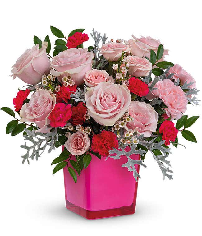 Pink roses, carnations, hot pink carnations, white waxflower, silver lace dusty miller, and huckleberry in a pink glass cube vase against a white background