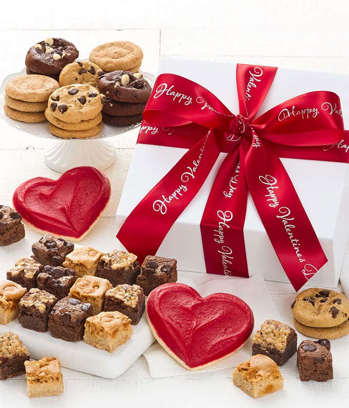 A white gift box with a red "Happy Valentine's Day" Ribbon with bite sized cookies laid out in front of the box along with two frosted heart cookies