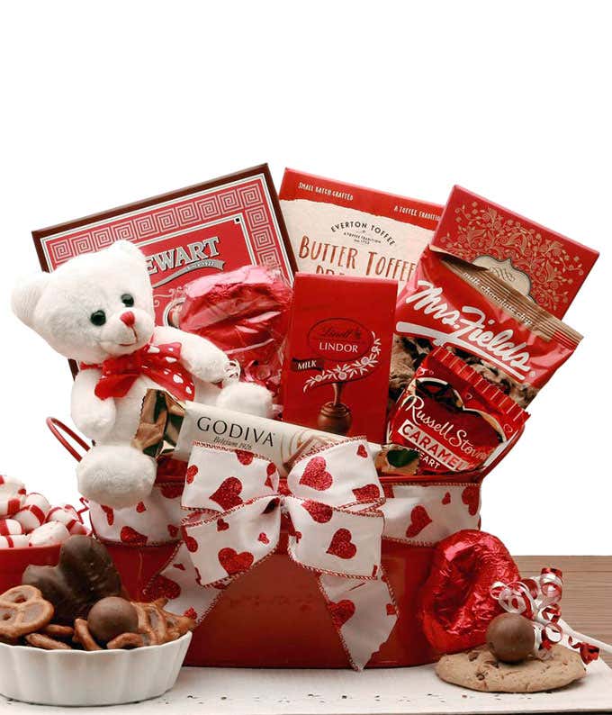 A red tin gift basket with white ribbon with red hearts. Inside, a whitet teddy bear, lindor chocolates, a Godiva chocolate bar, a caramel heart, and more items included can be seen